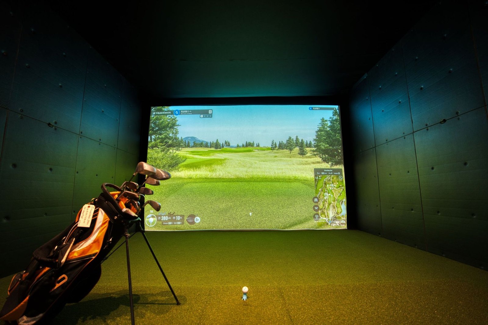 An indoor golf simulator bay with a screen showing a golf course, bar seating in the background